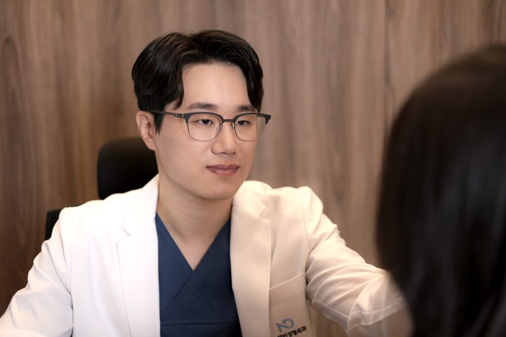 논현역 뉴데이성형외과 진료실에서 흰 가운을 입은 원장이 환자와 마주 앉아 상담하는 장면